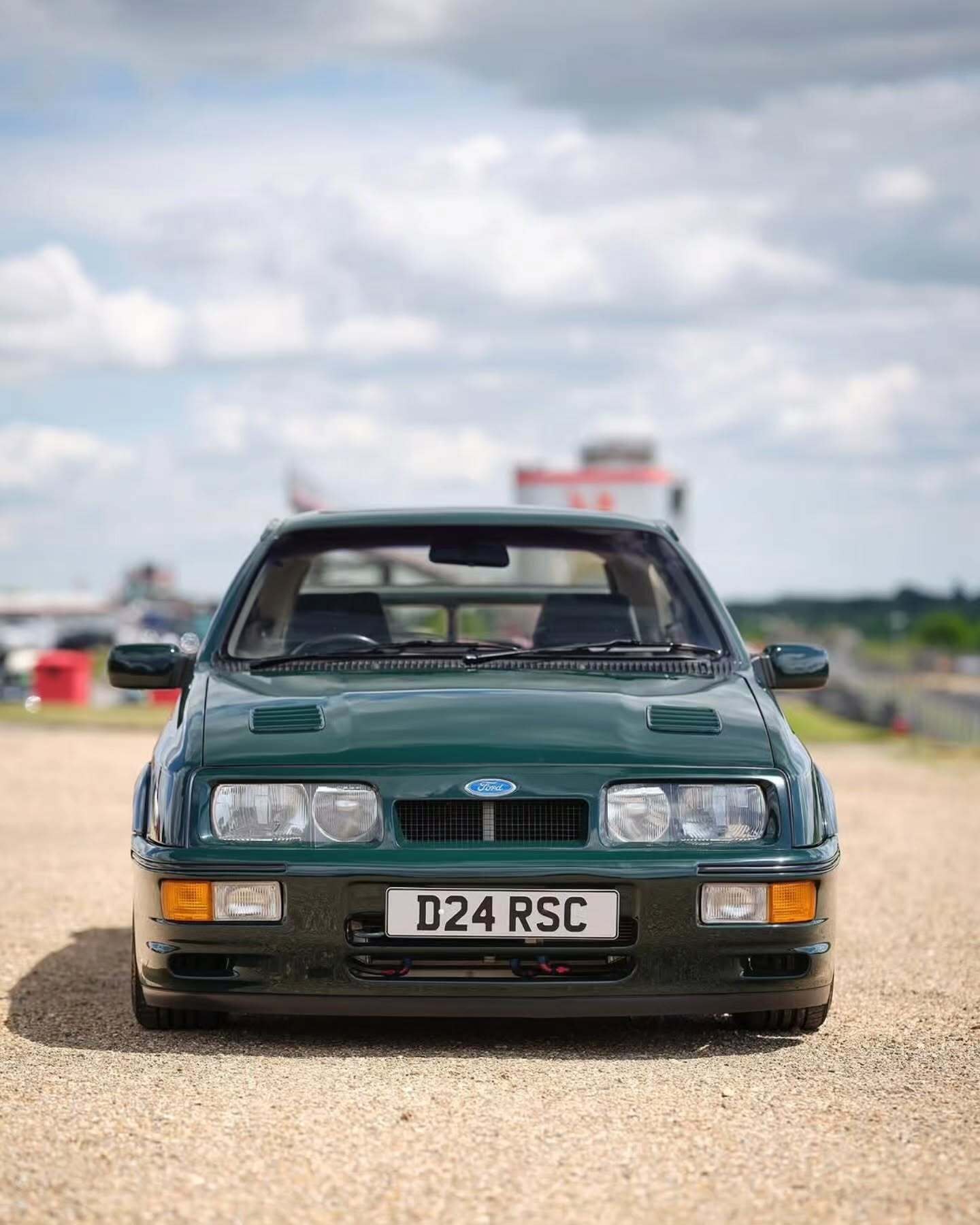 تصویر تمام‌قد و با زاویه باز از خودروی کلاسیک و اسپرت فورد سیرا کاسورث (Ford Sierra Cosworth) با رنگ تیره، که باله عظیم عقب و گلگیرهای برجسته آن در دهه ۱۹۸۰ میلادی آن را به یک نماد سرعت و سلطه بر پیست‌های مسابقه تبدیل کرد.