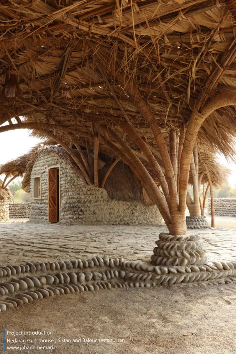 نمای خارجی و داخلی مهمانسرای ندارگ در روستای کهنانیکش، سیستان و بلوچستان، که ساختار نیمه‌باز، سقف چتری‌شکل با خرپاهای دست‌ساز و دیوارهای ضخیم گلی آن بر اساس بازتفسیر معماری بومی «کپر» و با هدف خنک‌سازی طبیعی و مشارکت جمعی اهالی طراحی و اجرا شده است.
