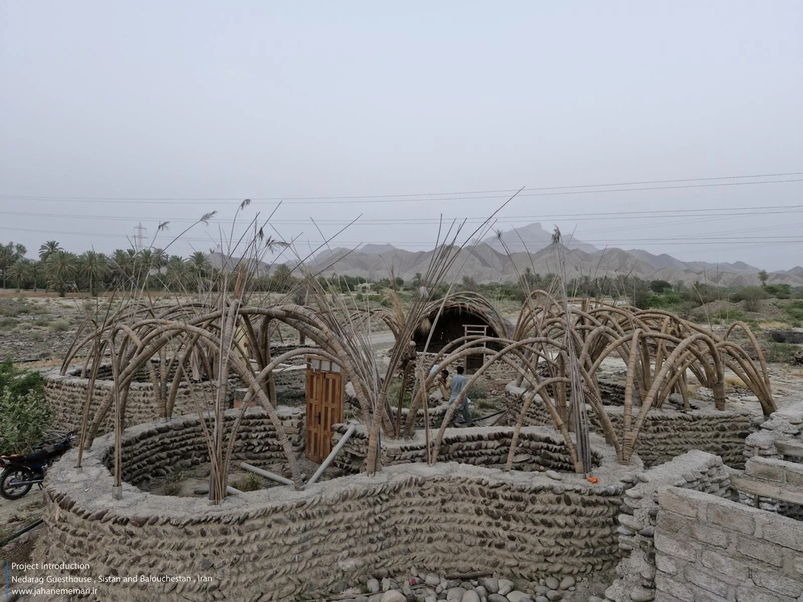 نمای خارجی و داخلی مهمانسرای ندارگ در روستای کهنانیکش، سیستان و بلوچستان، که ساختار نیمه‌باز، سقف چتری‌شکل با خرپاهای دست‌ساز و دیوارهای ضخیم گلی آن بر اساس بازتفسیر معماری بومی «کپر» و با هدف خنک‌سازی طبیعی و مشارکت جمعی اهالی طراحی و اجرا شده است.